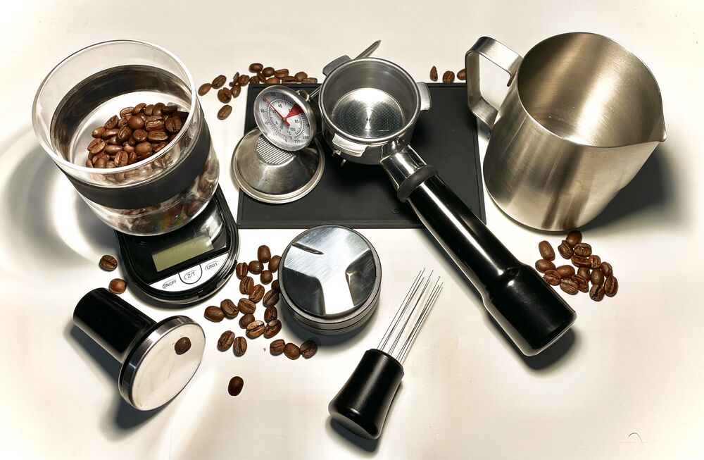 Espresso tools, a milk jug, a portafilter, a scale and coffee beans arranged on a pale surface.